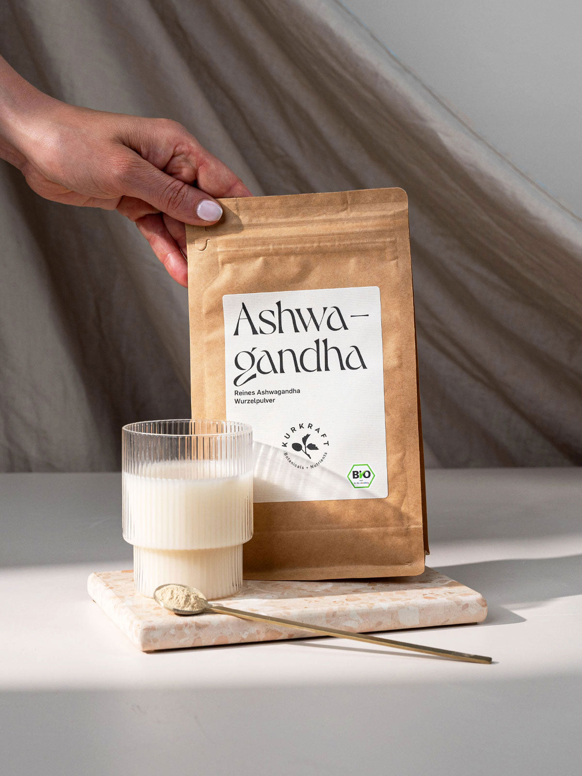 Ashwagandha Pulver von Kurkraft neben einem Glas Milch und Messlöffel auf hellem Untergrund