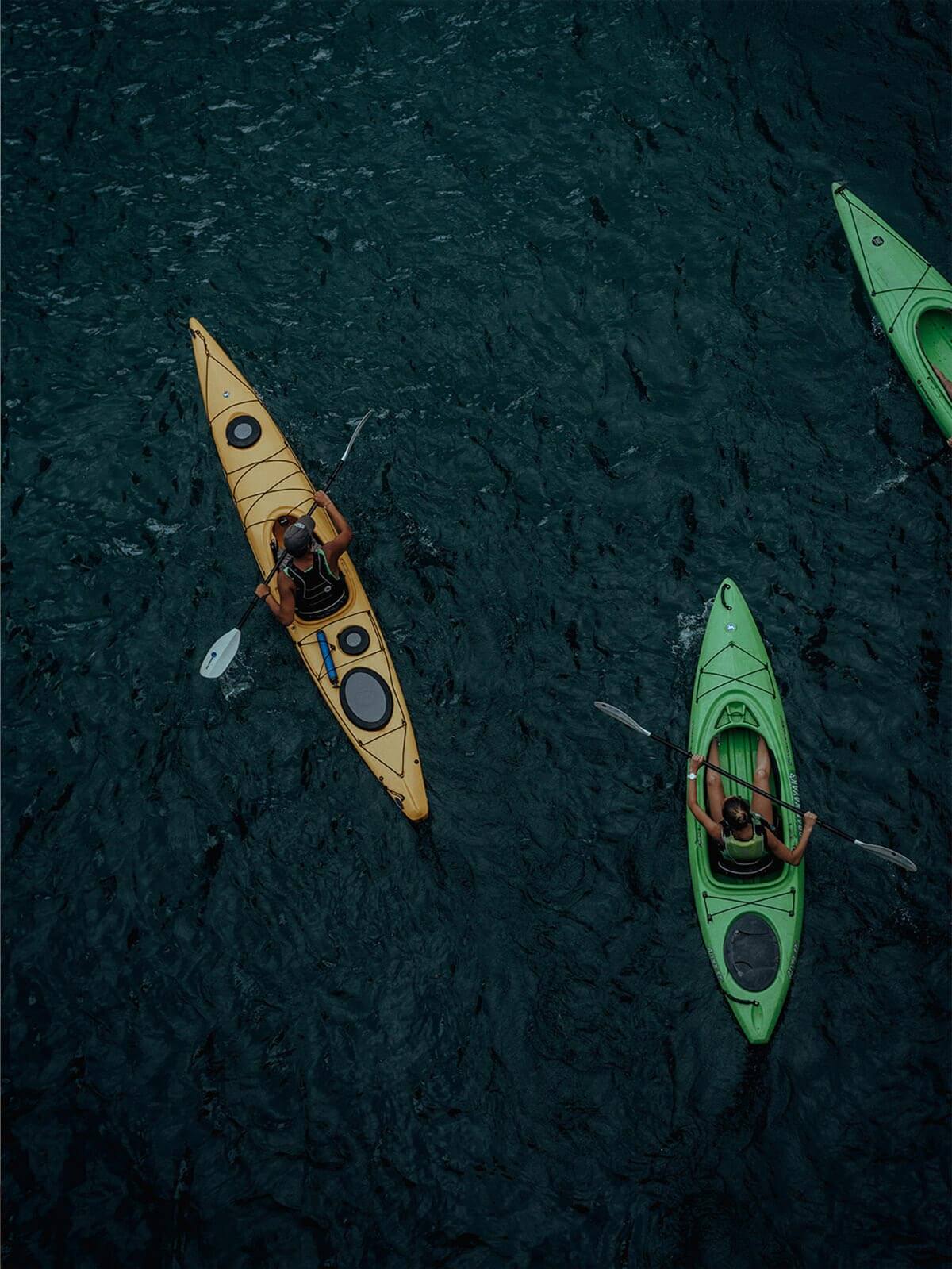 Zwei Personen paddeln in bunten Kajaks auf dunklem Wasser – Symbol für Bewegung, Energie und Gleichgewicht.