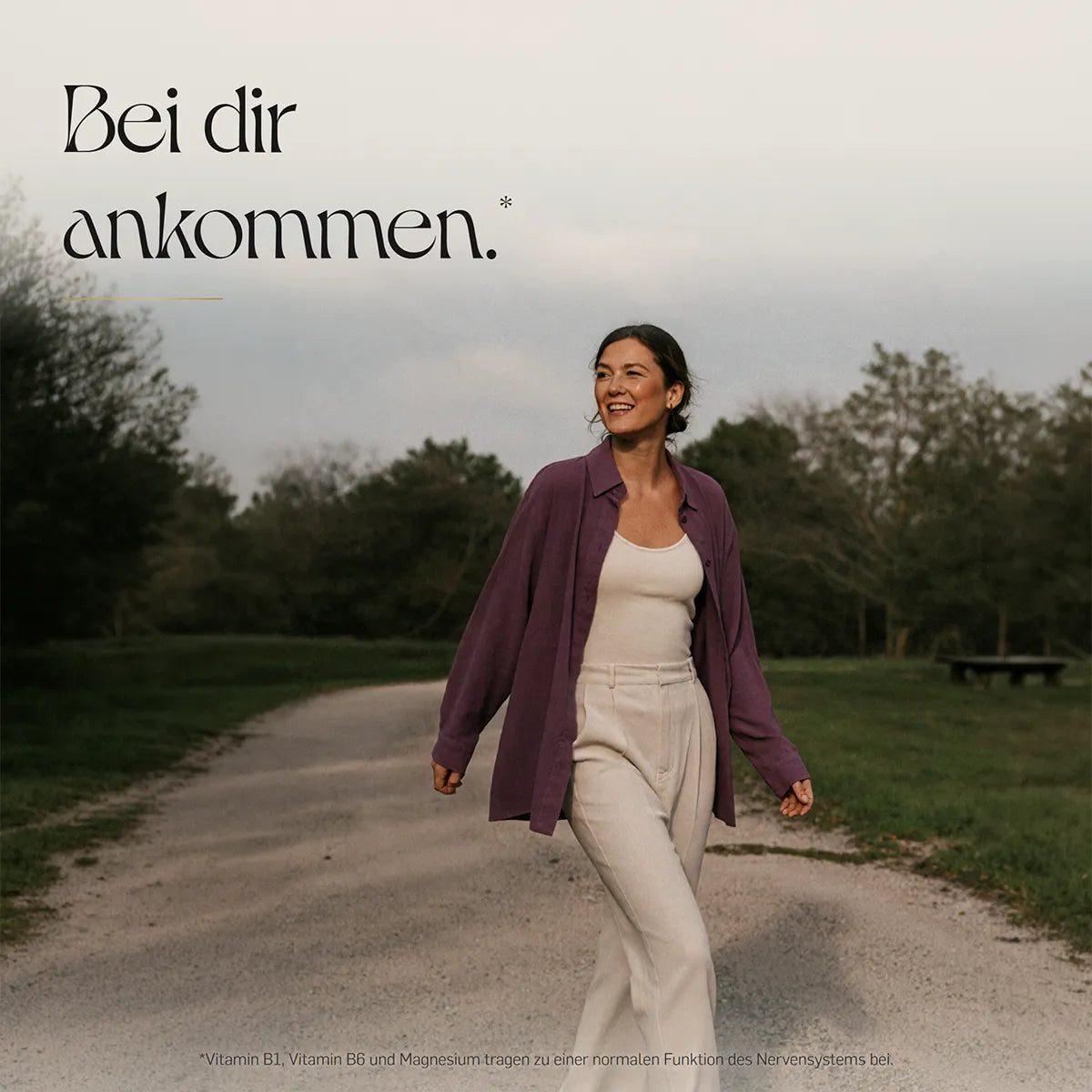 Bei dir ankommen – lächelnde Frau in fliederfarbener Bluse und heller Hose spaziert auf einem Kiesweg durch eine grüne Parklandschaft.