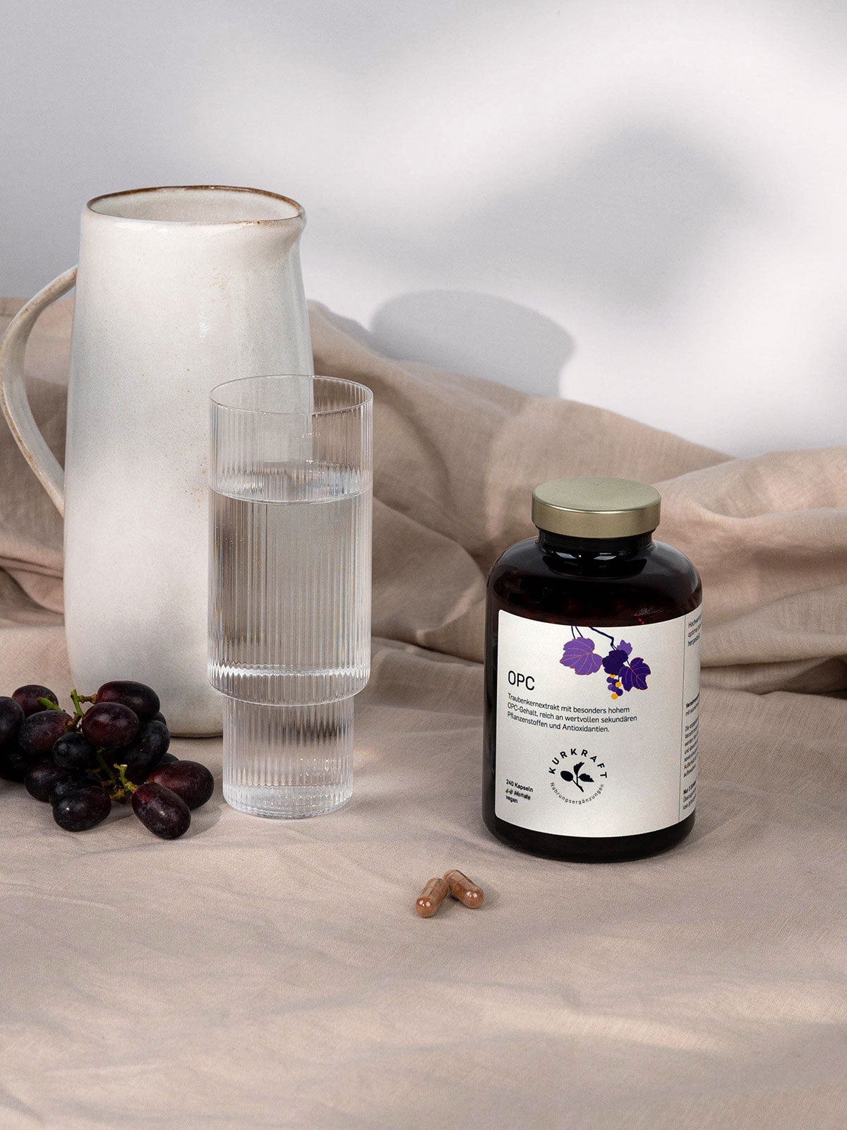 Kurkraft OPC Traubenkernextrakt Kapseln neben einem Glas Wasser, einer weißen Karaffe und frischen Trauben auf hellem Leinentuch.